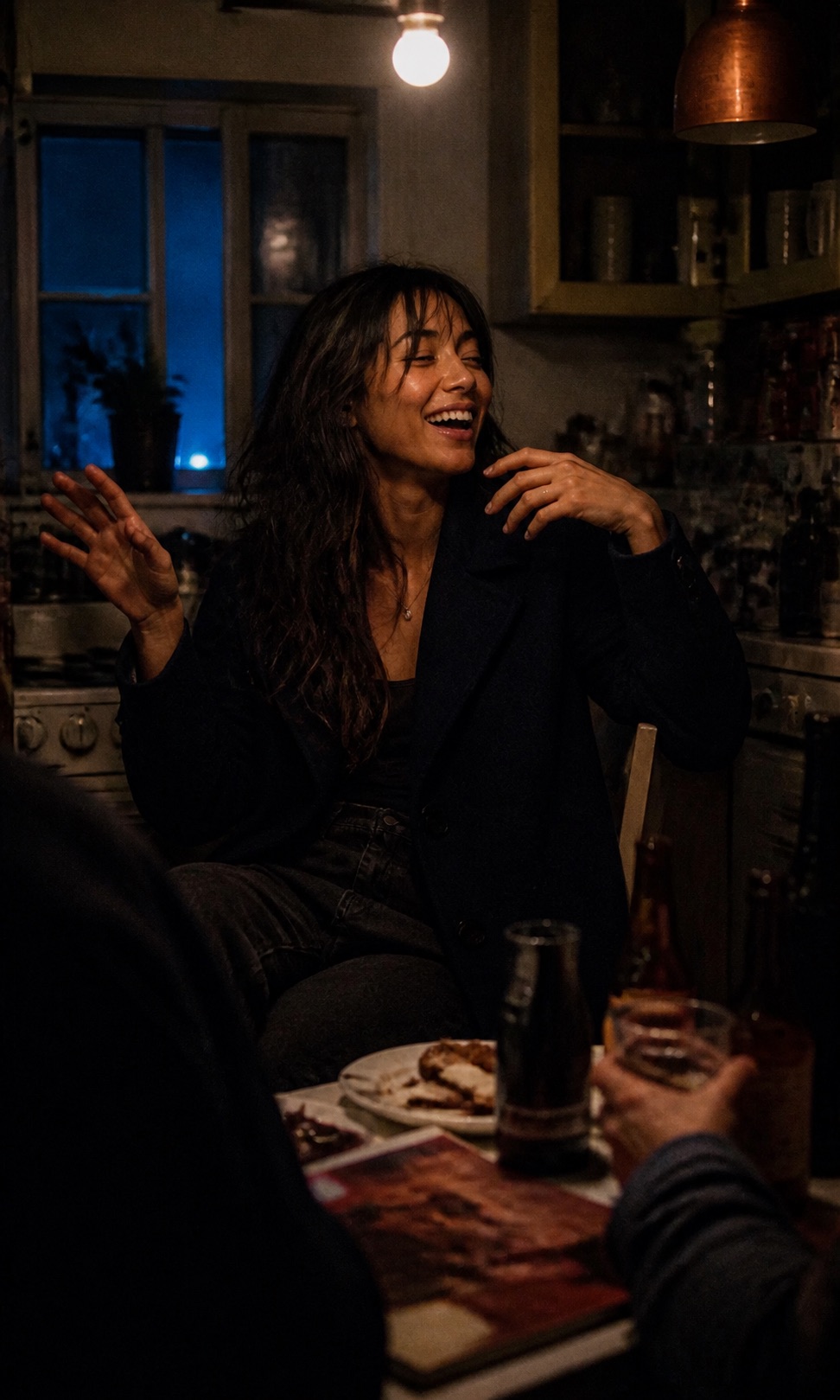 Nova Sinclair sitting sideways on a kitchen chair mid-laugh at one in the morning, hands mid-gesture, other people cropped and out of focus around the table.